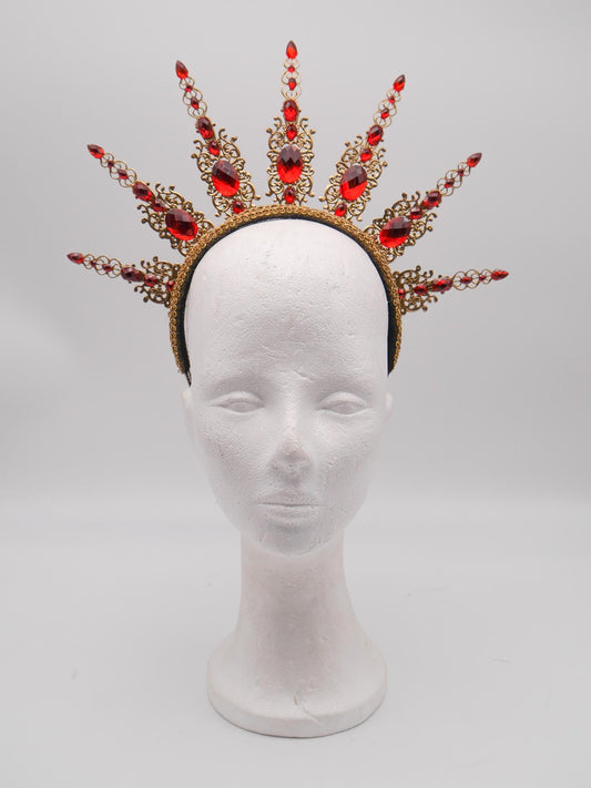 Könnte beinhalten: Ein goldfarbener Kronenaufsatz mit roten Edelsteinen und dekorativem Filigran. Die Krone sitzt auf einem schwarzen Stirnband und wird auf einem weißen Schaufensterkopf präsentiert. Die Edelsteine sind oval und tropfenförmig.
